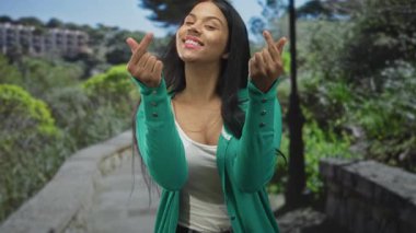 Latin kadın gülümsüyor iki eli taştan yolda, ağaçlarla ve tırabzanlarla çevrili yeşil parkta kalp hareketi yapıyor..