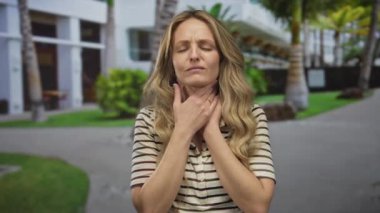 Güneşli bir günde dışarıda dikilen bir kadın boğazını tutuyor ve modern bir sokak ve yeşillik geçmişiyle pek iyi görünmüyor. Potansiyel sağlık sorunları olduğunu gösteriyor..