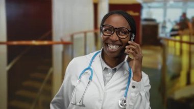 Doktor kadın otel lobisinde akıllı telefondan konuşuyor, gözlük ve laboratuvar önlüğü takıyor, steteskoplu, gülümsüyor ve mutlu bir şekilde el kol hareketi yapıyor..