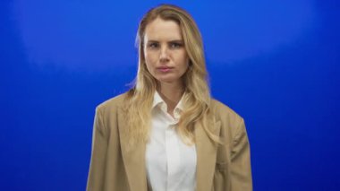 Kadın jestleri, elleri mavi arka planda durur, bej ceket ve beyaz gömlek giyer, temkinli veya ciddi bir ifadeyle uyarır..