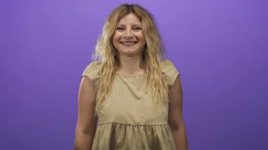 Günlük bej elbiseli, bol dalgalı bir kadın başını eğiyor ve stüdyoda kameraya dönük gülümsüyor..