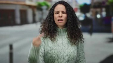 Açık havada, kıvırcık saçlı, süveter giyen, şehir arka planında el hareketleriyle öfke gösteren, şehir merkezinde hayal kırıklığını ifade eden bir kadın..