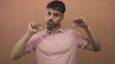 Pembe tişörtlü İspanyol adam başparmaklarını göğsüne doğru uzatıyor ve şeftali rengi stüdyo duvarına karşı dudaklarını büzüyor..