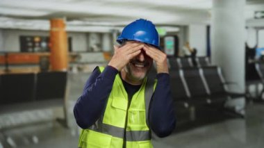 Havalimanı arka planında mavi kasklı ve sarı yelekli bir adam gülerek neşeli duygular ve bulanık oturma yeriyle günlük kıyafetlerini gösteriyor..