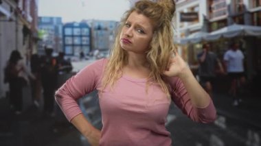 Sarışın genç kadın saçlarıyla oynuyor ve kalabalık bir caddede dükkanlar ve kafelerle el ele dinleniyor, binaların yanında duruyor; sıkıntı.