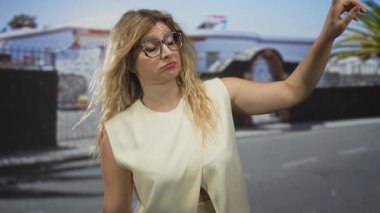 Büyük gözlüklü kadın banliyö caddesinin girişinde parmağını gösterirken, kolu avuç içi kadar açık; şüphecilik.