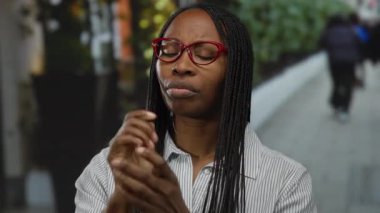 Kentsel sokakta bilek ağrısı çeken kıvırcık saçlı ve gözlüklü bir kadın, açık havada rahatsızlığını gösteriyor, şehir arka planında günlük gömleğini giymiş bir kadın..