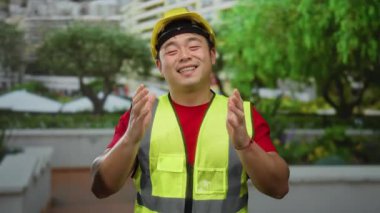 Güvenlik yeleği ve kasklı gülümseyen Asyalı adam arka planda yemyeşil yemyeşil bir caddede güvenle dikiliyor..