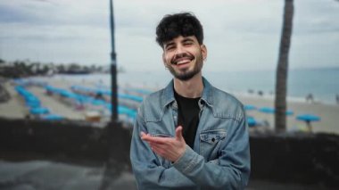 Kumsalda gülümseyen genç bir adam bulutlu bir gökyüzünün altında kot ceket giyiyor, deniz kenarında bir atmosferde boş zamanlarını ve mutluluğu yakalıyor..