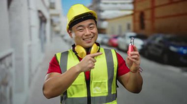 Güler yüzlü Asyalı adam emniyet yeleği giyiyor ve kentsel caddedeki elektronik cihaza nokta koyuyor. Arka planda arabaları var. Şehir temalı açık hava portresi çizimleri yapıyor.