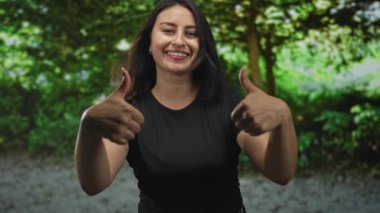 Genç İspanyol kadın gülümsüyor ve yeşil ağaçlar ve yapraklar arasında ormanda iki parmağını da gösteriyor; sevinç onayı.