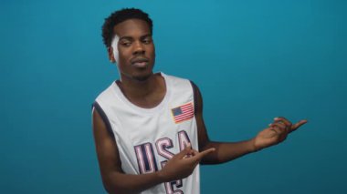 Beyazlı Amerikan basketbol forması giyen adam teal stüdyosunda gülümserken bayrak yamasını işaret ediyor..