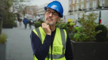 Yansıtıcı yelek ve sert şapka giyen olgun bir inşaat işçisi bir şehir caddesinde düşünceli bir şekilde duruyor, güvenlik ve profesyonellik üzerine yoğunlaşarak kentsel alana karışıyor..