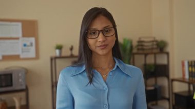 Ofis odasında gülümseyen genç bir kadın. Gözlüklü, mavi tişörtlü, ilan tahtası ve arka planda saksı bitkisi olan bir kadın. Profesyonel bir ortam yaratıyor.