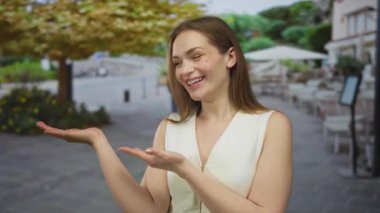 Beyaz kolsuz bluzlu genç bir kadın güneşli caddede heyecanlı bir şekilde el kol hareketi yaparak avuçlarını uzatıyor; dostça bir davet..