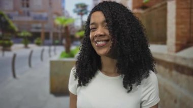 Sokaktaki tuğla binanın önünde dişlerini ve kıvırcık saçlarını gösteren Afrikalı Amerikalı kadın gülümsüyor. İyimserlik..