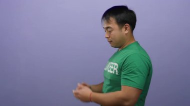 Yeşil tişörtlü gülümseyen Asyalı adam, mor arkaplan karşısında ellerini boş tutuyor. İletişim ve etkileşimi ifade ediyor.