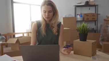 Yeni evde dizüstü bilgisayar kullanan bir kadın. Elinde kredi kartı olan karton kutularla dolu. Modern yaşam tarzını ifade eden ve internetten alışveriş yapan bir kadın..