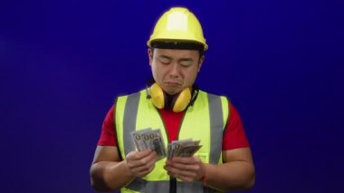 Güvenlik yeleği ve kasketli genç adam mavi arka planda duruyor. Düşünceli bir ifadeyle dolarları tutuyor. Finansal kararları ya da inşaat sektörünü temsil ediyor..