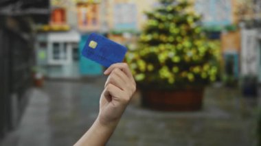 Canlı bir şehir sokağında elinde kredi kartı tutan beyaz bir adam. Arka planı bulanık. Renkli binalar ve bir ağaç var..