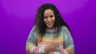 Canlı mor duvarın üzerinden gökkuşağı süveteri içinde gülümseyen bir kadın neşeli bir ruh hali ve kıvırcık saçlı izole bir arkaplan tasvir ediyor..