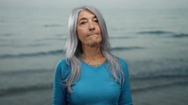 Deniz kenarında dikilirken gri saçlı yaşlı bir kadın okyanusun dingin zeminine mavi bir kazak giyip dalgalar savuruyor..