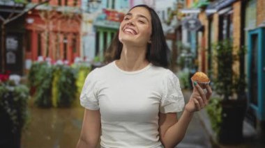 Kek tutarken gülümseyen ve yukarı bakan bir kadın işlek caddede parlak güneş ışığı altında beyaz gömlek giyerek; neşe.