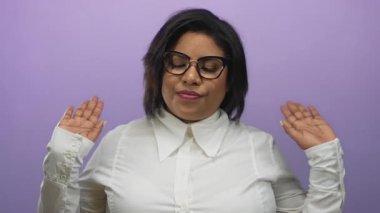 Kendinden emin, iri yarı İspanyol bir kadın canlı mor bir duvara el kaldırarak el kol hareketi yapıyor, gözlük takıyor ve beyaz gömlek giyiyor..