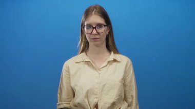 İşaret parmağıyla işaret parmağıyla işaret eden bej gömlekli ve gözlüklü bir kadın, sağlam mavi bir arka planı olan bir stüdyoda..