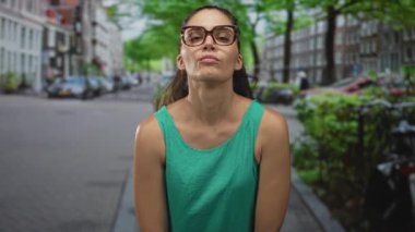 Kadın kollarını havaya kaldırır ve yeşil bir kolsuz tişörtle şehir caddesinde ellerini karnına bastırır, gözlük görünür, özgüven odaklıdır..