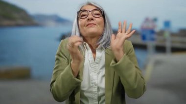 Gri saçlı ve gözlüklü bir kadın sahilde sevinçle duruyor, mutluluk ve sükunet yayıyor, yeşil bir ceket giyiyor ve doğayı kucaklıyor..