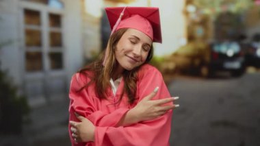 Pembe mezuniyet elbisesi ve kepi giymiş bir kadın güneşli bir şehir arka planında güvenle gülümsüyor, akademik başarıyı dışarıda kutluyor..