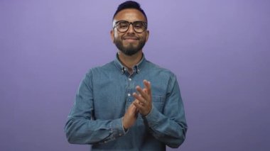 Adam el çırpıyor ve stüdyoda gülümsüyor kot gömlek ve mor arka planlı gözlük takıyor; sevinç kutlaması.