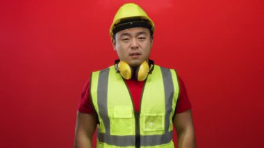 Sarı güvenlik yeleği giyen ve kasketli genç adam canlı kırmızı bir arka planda duruyor. İnşaata hazırlık ve profesyonelliği somutlaştırıyor..