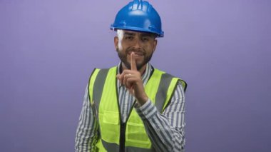 Mor stüdyoda mavi kasketli ve yüksek görünürlüklü yeleği, parmak dudağa hareket ve çizgili gömleği olan adam mühendisi, sessizlik..