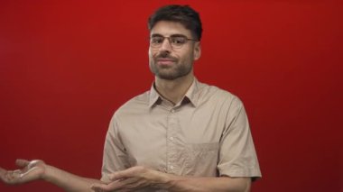 Kırmızı stüdyoda bej gömlekli ve gözlüklü bir adam açık avuç içi jesti ve ince bir gülümseme; merak.