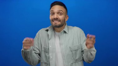 Genç İspanyol bir adam neşeli bir gülümseme ile canlı mavi bir arka plana karşı omuz silkiyor, rahat bir gömlek giyiyor, mizah duygusu ve oyunculuk taşıyor..