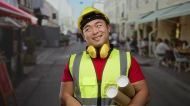 Güvenlik teçhizatı takan gülümseyen adam arka planda kafeleri olan bir şehir sokağının planlarını tutuyor..