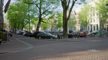 Güneşli bir günde yeşil ağaçlarla, park edilmiş arabalarla ve bisikletlerle çözünmüş arka plandaki bulanık sokak manzarası huzurlu bir şehir atmosferi yaratıyor..