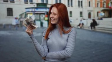 Şehir caddesinde İsveçli banknotlarla gülümseyen bir kadın, arka planda tarihi binalar bulunan açık şehir ortamında kızıl saçlı güzelliği sergiliyor..