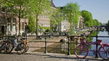 Amsterdam 'da bisiklet, yeşillik ve tarihi binalarla bulanık kanal manzarası berrak mavi bir gökyüzünün altında sakin su yolunu kaplıyor..