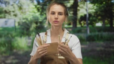 Yeşil bir park ortamında elinde fırça tutan kadın ressam, önlük giyiyor, açık hava resim faaliyetlerine hazırlanırken yüzünde odaklanmış bir ifade var..