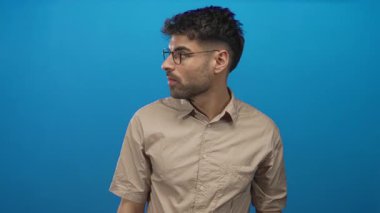 Ağzı açık ve kafası yana dönmüş bir adam mavi stüdyo duvarına karşı gözlüklü ve bej gömlekli; sürpriz.