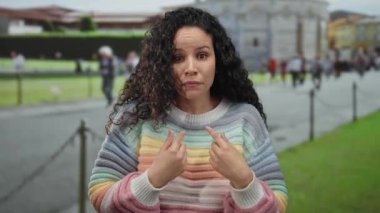 Renkli süveter giyen genç İspanyol kadın şaşkınlığını belirtirken, İtalya 'da bulanık kalabalıkla birlikte açık hava turist seti olan Pisa Kulesi' nin arka planına işaret ediyor..