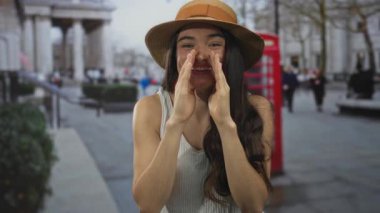 Hasır şapkalı genç bir kadın kırmızı telefon kulübesinin yanındaki caddede bağırmak için elleri ağızlarının etrafına dolanıyor; heyecan enerjisi.