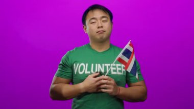 Yeşil gönüllü Asyalı genç adam pembe arka planda Tayland bayrağı tutuyor.