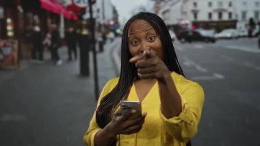 Sarı tişörtlü bir kadın, bulanık arka plan ve çeşitli yayaları olan şehir sokak ortamında telefonu işaret ediyor ve tutuyor..