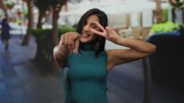 Açık havada gülümseyen, barış işareti yapan ve gündüz vakti şehir caddesinde kameraya bakan neşeli ifade ve pozitif enerji sergileyen bir kadın..