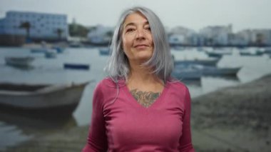 Kır saçlı yaşlı kadın sahilde teknelerin yanında şakacı kulak hareketleri yapıyor, uzak binaları olan sakin bir deniz kıyısına karşı neşeli kişiliğini sergiliyor..