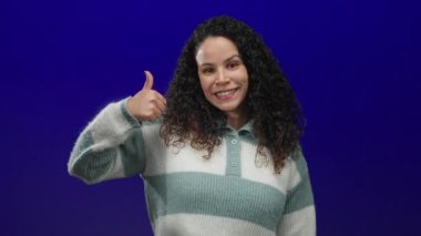 Kıvırcık saçlı, çizgili süveter giyen, onay ve pozitiflik gösteren, izole edilmiş mavi arka planı onaylayan gülümseyen kadın..